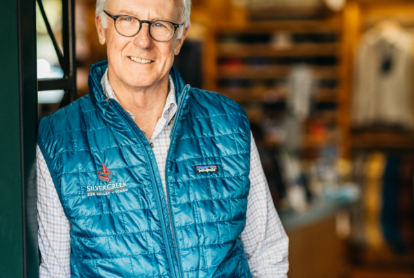 An older man with glasses and gray hair, wearing a blue quilted vest over a checkered shirt, stands indoors and smiles at the camera; shelves and clothes are blurred in the background.