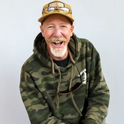 A smiling man with a mustache and beard wears a camouflage hoodie, tan cap with a patch, and sunglasses hanging from his neck, sitting against a plain light background.