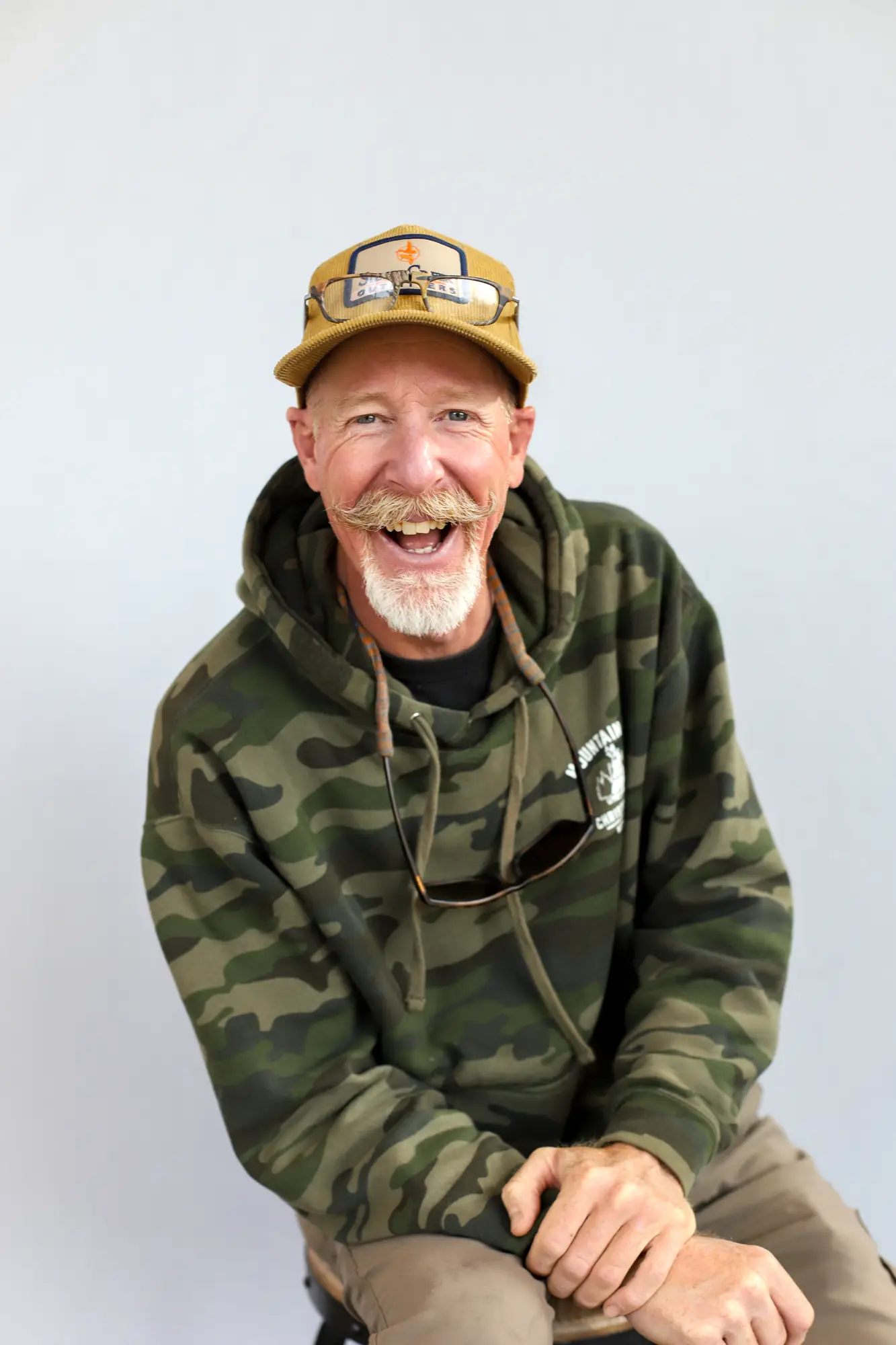 A smiling man with a mustache and beard wears a camouflage hoodie, tan cap with a patch, and sunglasses hanging from his neck, sitting against a plain light background.