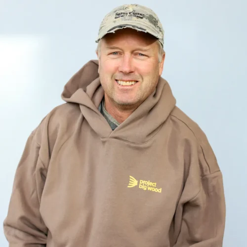 A man wearing a tan Project Big Wood hoodie and a light-colored baseball cap smiles at the camera, embodying the spirit of Sun Valley fly fishing guides against a plain light background.
