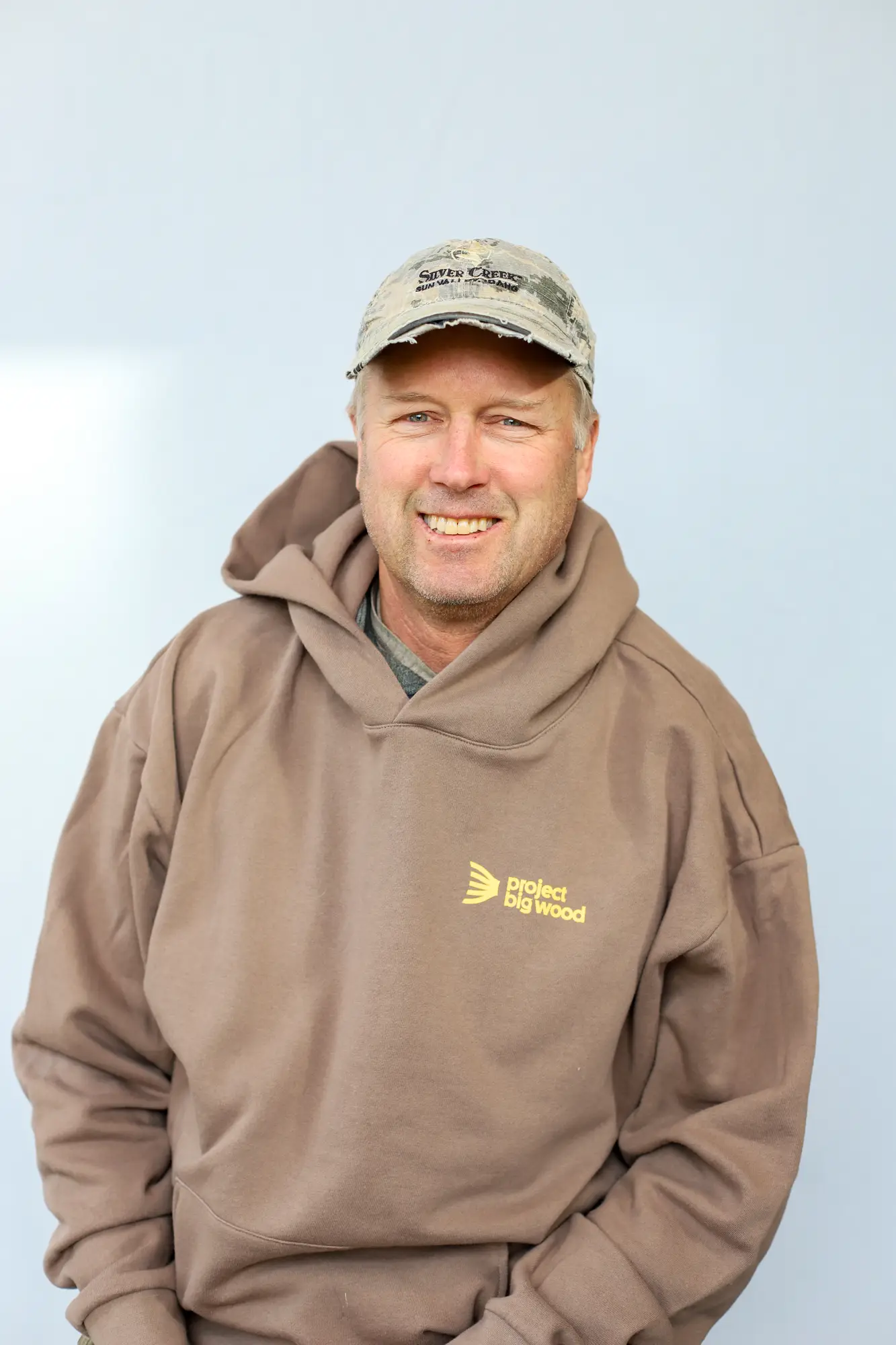 A man wearing a tan Project Big Wood hoodie and a light-colored baseball cap smiles at the camera, embodying the spirit of Sun Valley fly fishing guides against a plain light background.
