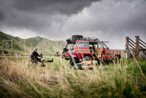 A person sits on a camping chair near a red SUV with open doors, parked on grassy land under a cloudy sky. Camping gear is spread out, and green hills are visible in the background.