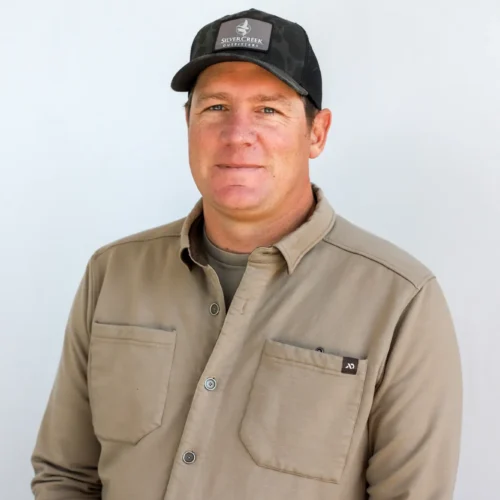 John Grigsby, a Fly Fishing Guide from Sun Valley, Idaho, stands against a plain light background in a black baseball cap and beige button-up shirt, facing the camera with a neutral expression.