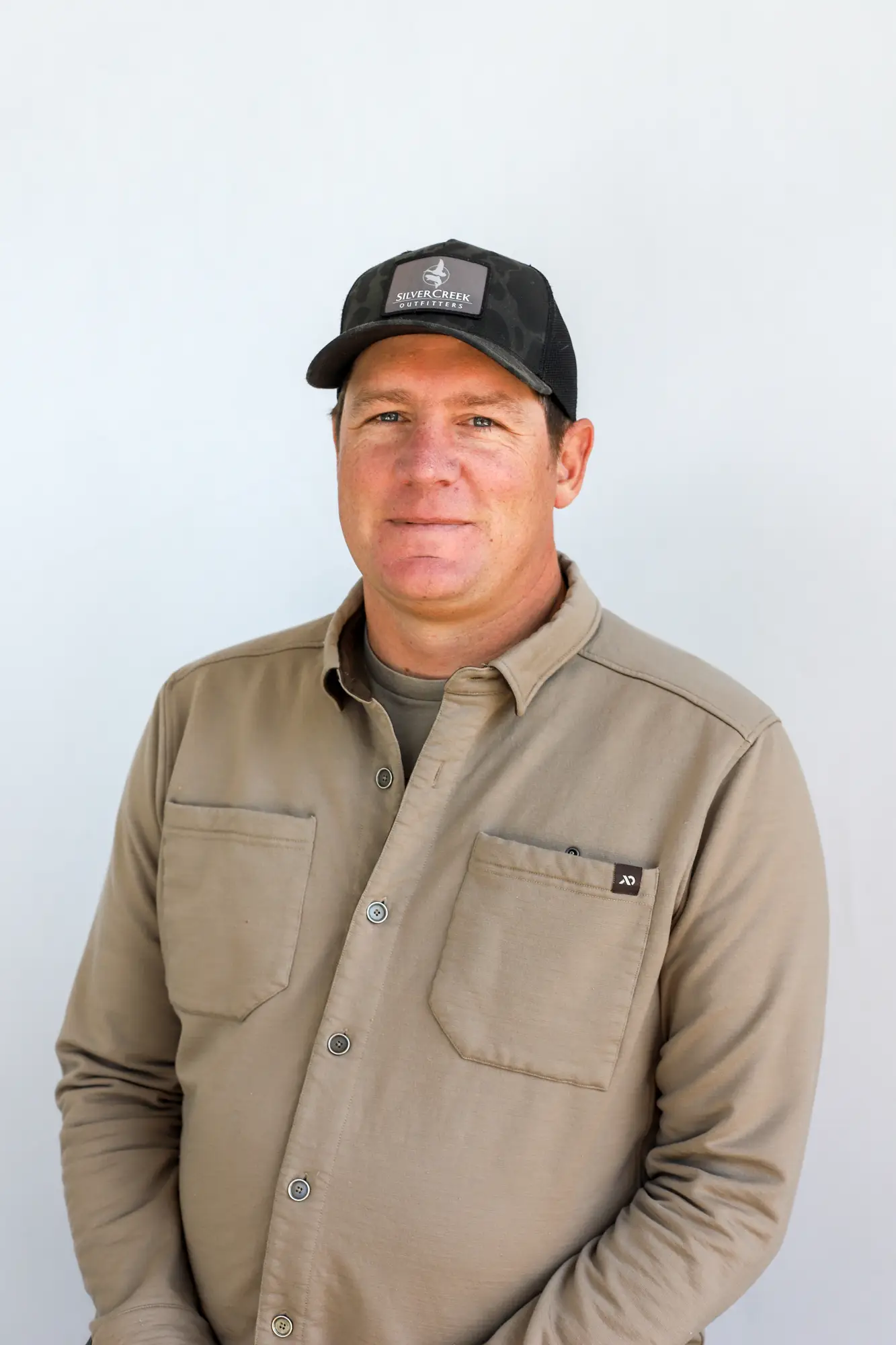 John Grigsby, a Fly Fishing Guide from Sun Valley, Idaho, stands against a plain light background in a black baseball cap and beige button-up shirt, facing the camera with a neutral expression.