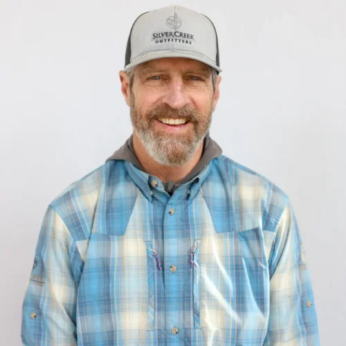 A man with a brown beard and mustache smiles at the camera. Certified Casting Instructor Scott Connell, a Fly Fishing guide, is wearing a gray cap, blue plaid shirt, and gray hooded garment, standing before a plain light background.