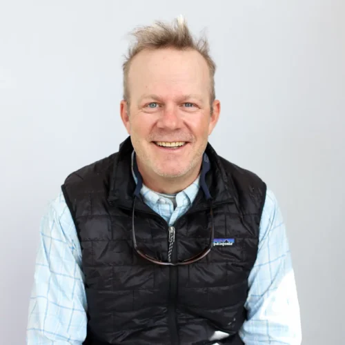 A smiling man with short, messy blond hair wearing a light blue plaid shirt, black quilted vest, and sunglasses hanging from his neck sits in front of a plain gray background—ready for his next Sun Valley Fly Fishing Guide adventure.