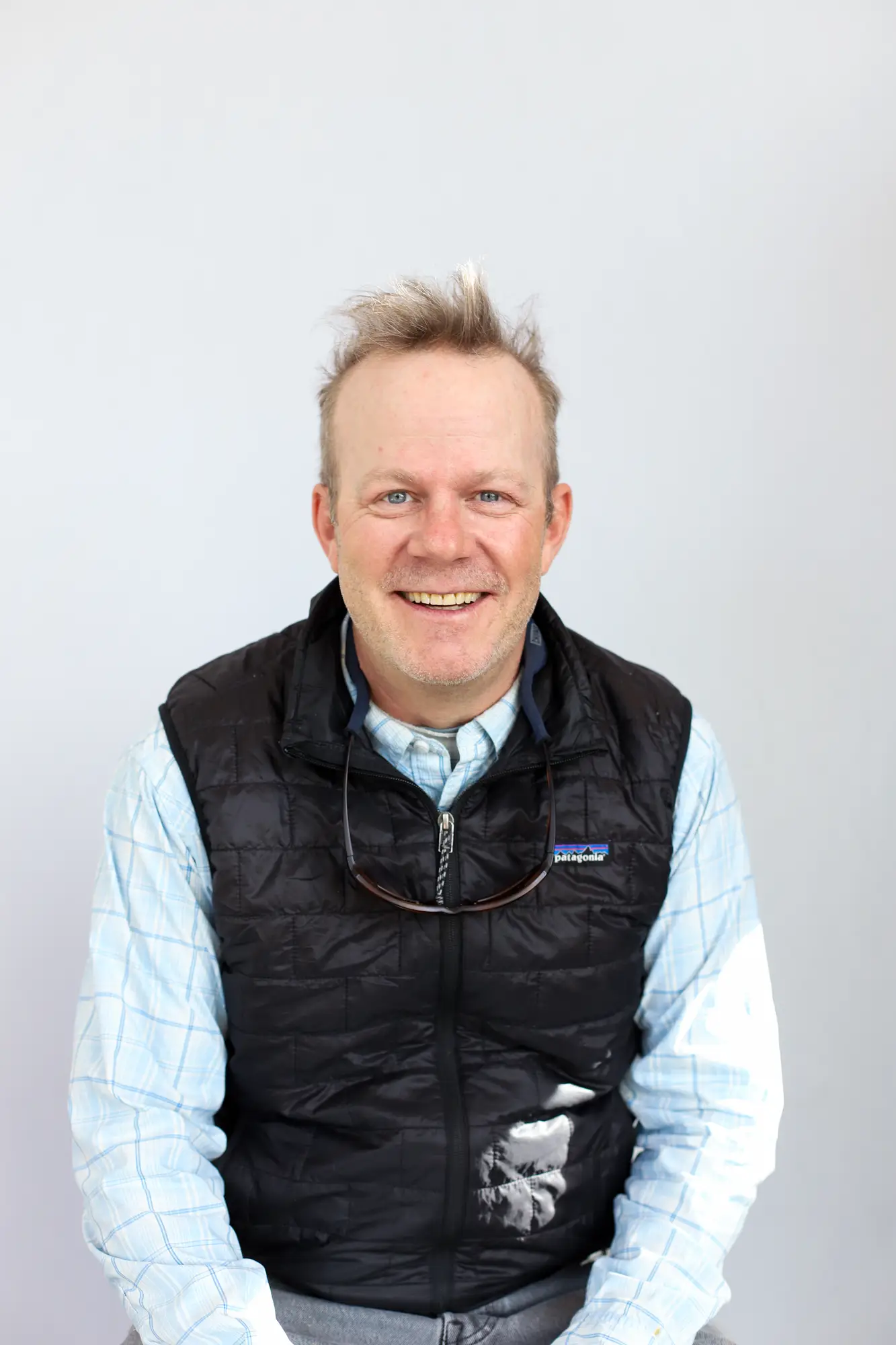 A smiling man with short, messy blond hair wearing a light blue plaid shirt, black quilted vest, and sunglasses hanging from his neck sits in front of a plain gray background—ready for his next Sun Valley Fly Fishing Guide adventure.
