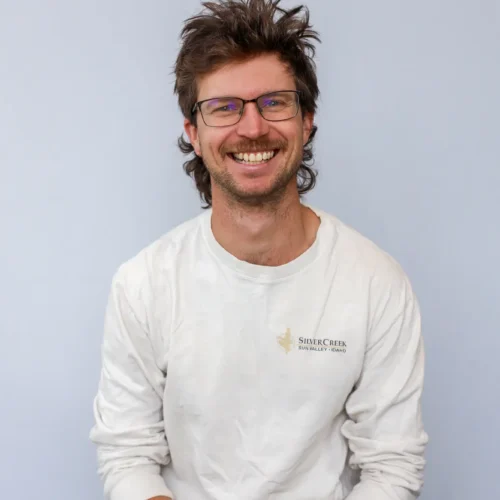A man with glasses and tousled hair, wearing a white long-sleeve shirt with a Fly Fishing Guide logo and dark jeans, sits against a plain light gray background, smiling broadly at the camera.