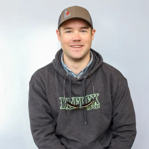 A man wearing a gray hoodie with Reflex Ski Poles text, a gray baseball cap, and sunglasses hanging from his collar sits against a plain light background, smiling at the camera. This is Matt O'Connor, an FFF certified casting instructor.