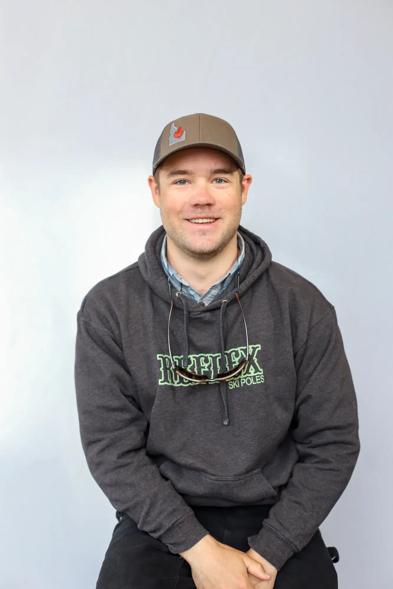 A man wearing a gray hoodie with Reflex Ski Poles text, a gray baseball cap, and sunglasses hanging from his collar sits against a plain light background, smiling at the camera. This is Matt O'Connor, an FFF certified casting instructor.