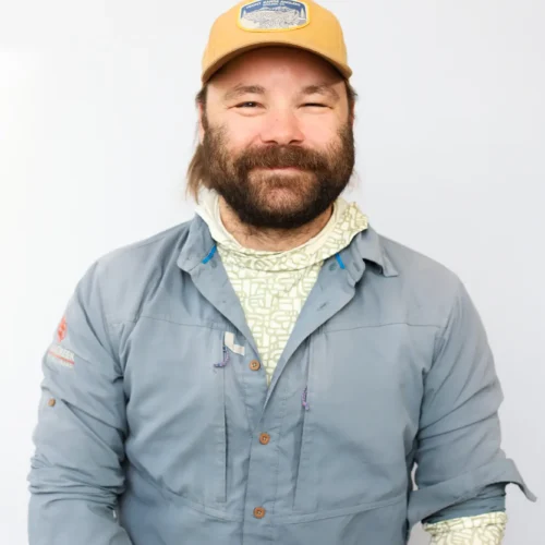 A man with a beard and mustache wearing a tan cap, light blue button-up shirt, and patterned neck gaiter smiles against a plain light background.