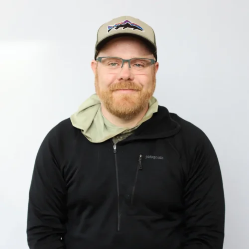 A bearded man wearing glasses, a beige cap with a mountain logo, and a black zip-up jacket sits against a plain white background, smiling softly at the camera—Certified Casting Instructor Lewis Hunt.