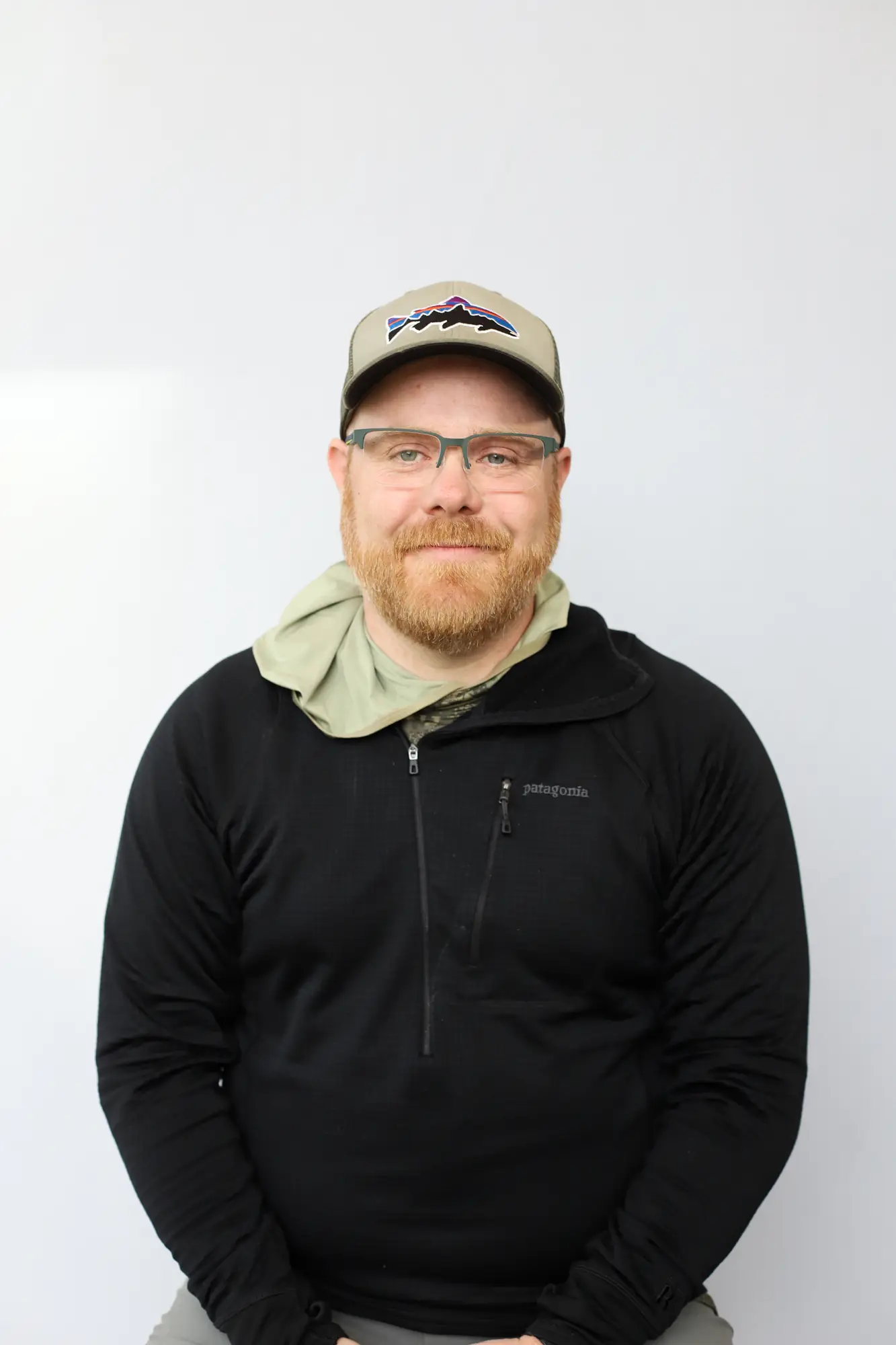 A bearded man wearing glasses, a beige cap with a mountain logo, and a black zip-up jacket sits against a plain white background, smiling softly at the camera—Certified Casting Instructor Lewis Hunt.