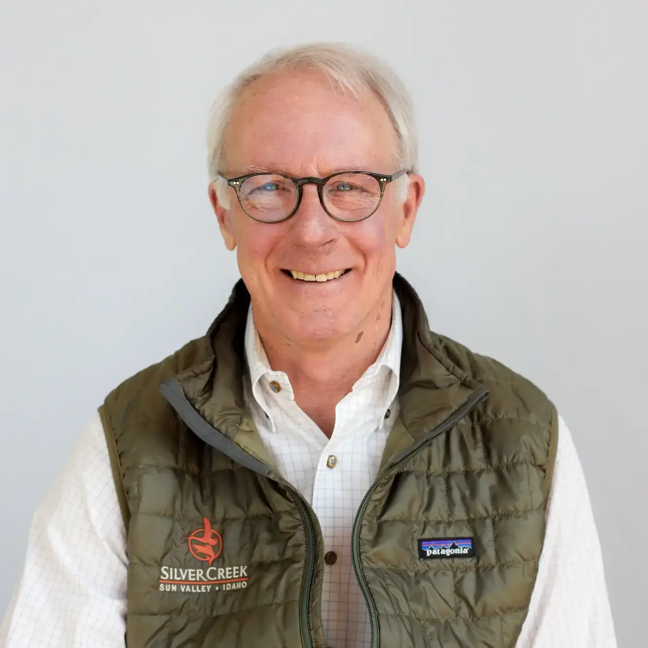 A man with white hair and glasses smiles at the camera. He is wearing a white shirt and a green vest with “Silvercreek Sun Valley Idaho” and “Patagonia” logos, identifying him as Terry Ring, the Owner. The background is plain and light-colored.