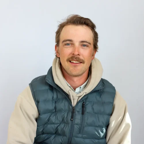 A man with light brown hair and a mustache, wearing a beige hoodie and a teal puffer vest, sits and smiles against a plain background—ready to share tips as your trusted Sun Valley Fly Fishing Guide.