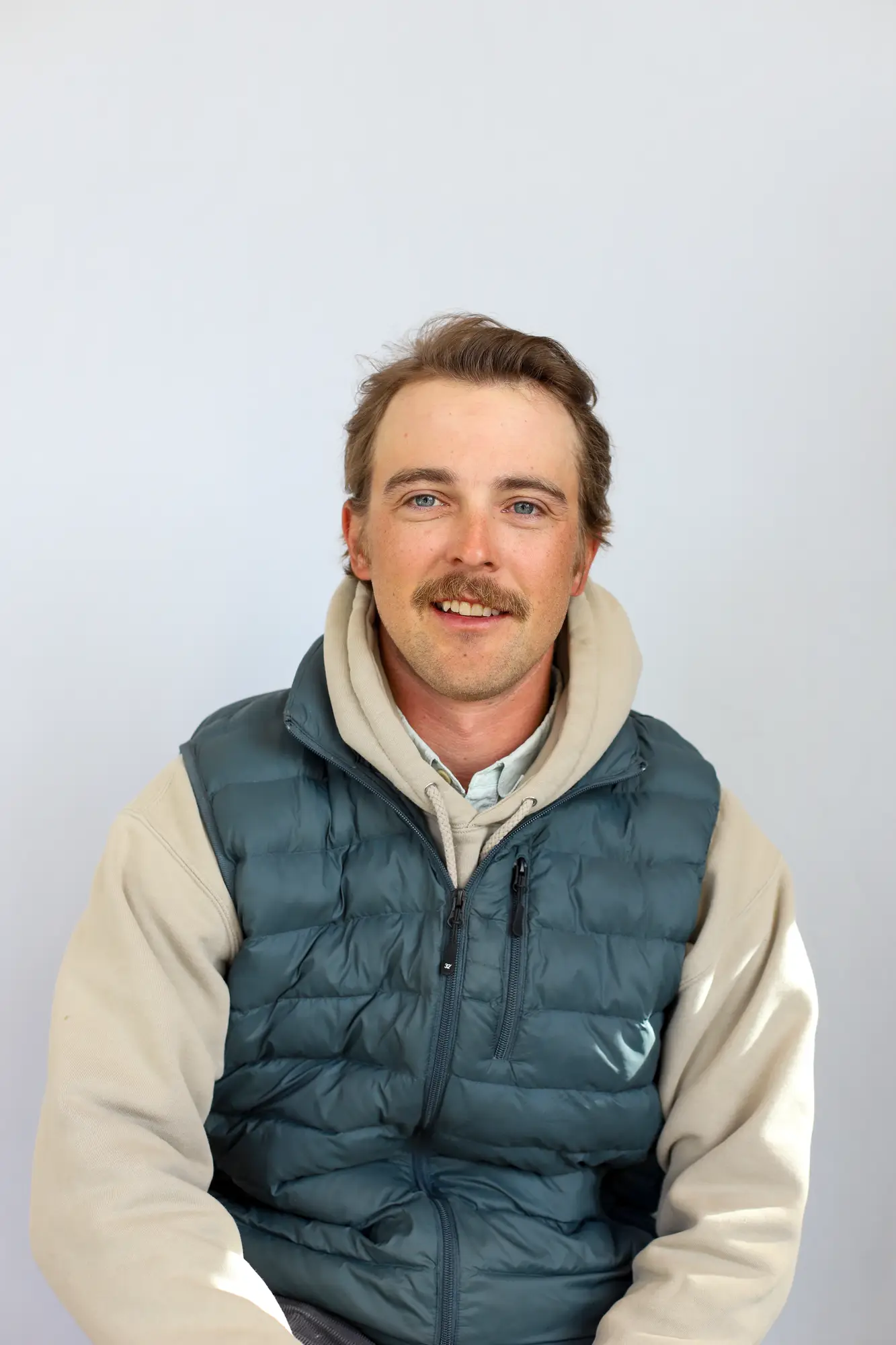 A man with light brown hair and a mustache, wearing a beige hoodie and a teal puffer vest, sits and smiles against a plain background—ready to share tips as your trusted Sun Valley Fly Fishing Guide.