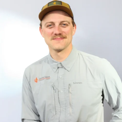 A man with a light brown mustache, dressed in a brown and green cap and a light gray long-sleeve shirt, smiles warmly as he sits in front of a plain white background—he looks every bit the friendly fly fishing guide.