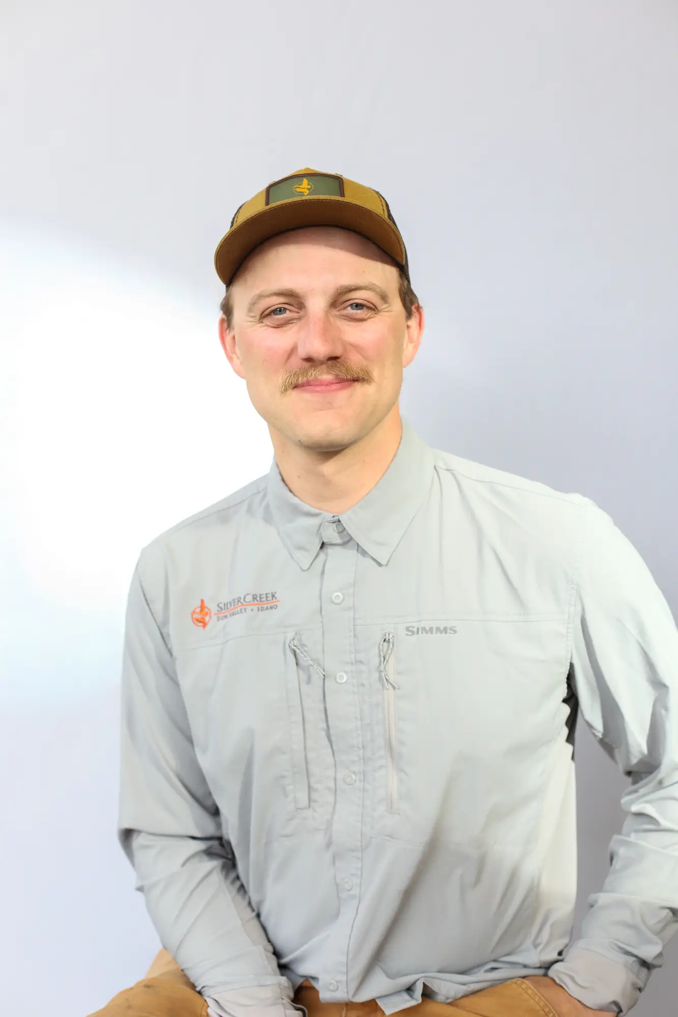 A man with a light brown mustache, dressed in a brown and green cap and a light gray long-sleeve shirt, smiles warmly as he sits in front of a plain white background—he looks every bit the friendly fly fishing guide.