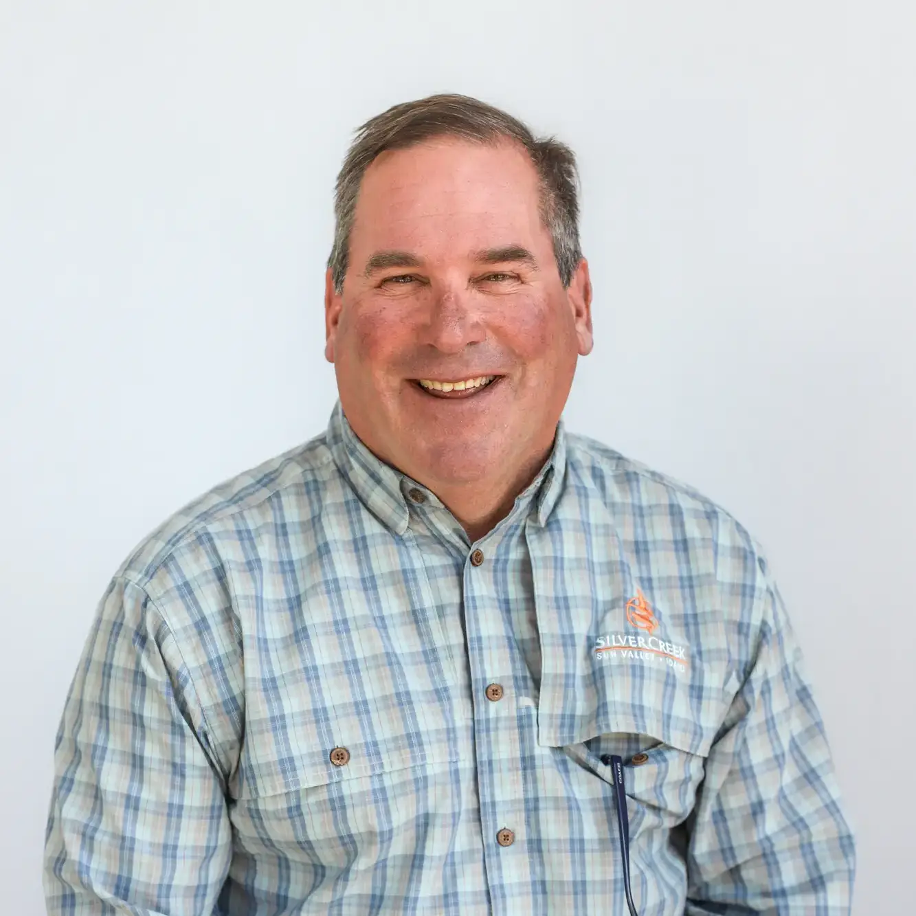 A middle-aged man with short brown hair smiles at the camera. He is wearing a blue and white plaid button-up shirt with a Silver Creek Outfitters logo, standing against a plain light background. He looks every bit the friendly Retail Associate.