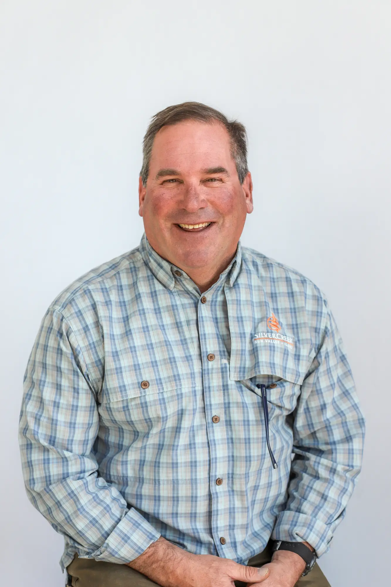 A middle-aged man with short brown hair, wearing a blue and white plaid Silver Creek Outfitters shirt with a logo on the chest, sits and smiles in front of a plain light background.