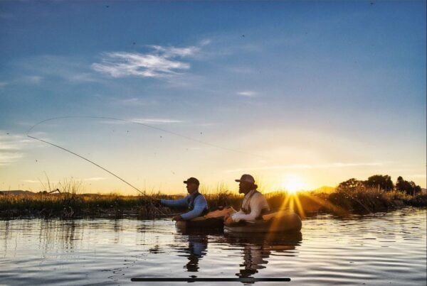 Two people sit in a small boat on calm water, enjoying spring fly fishing Idaho at sunrise. The sun is low on the horizon, casting warm light and reflections as clear skies and grass line the water’s edge.