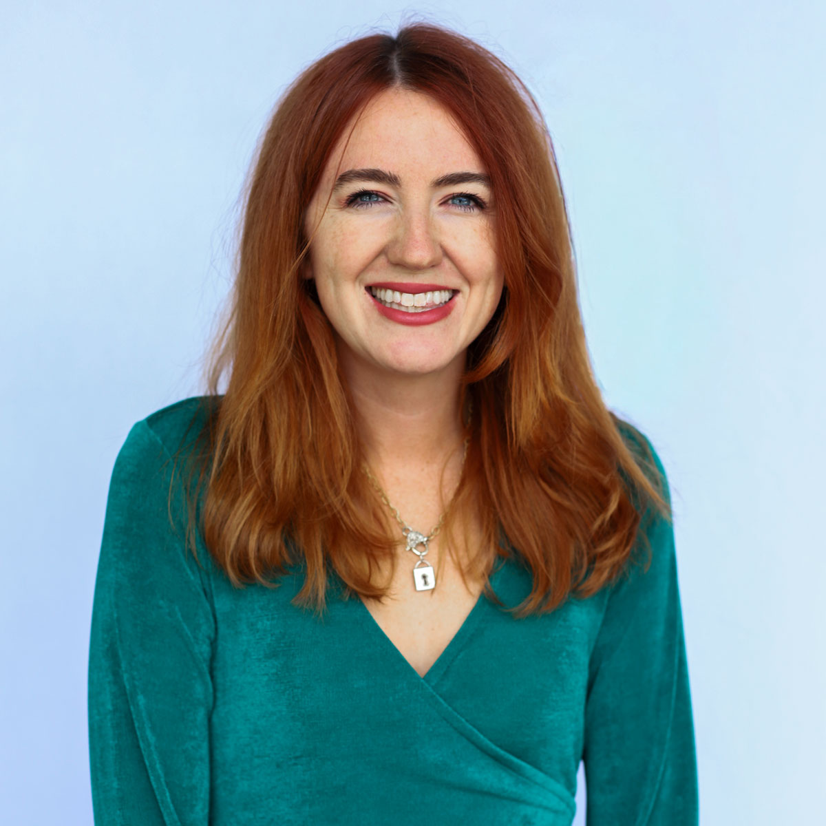 Madeline Funk, a Women's Apparel Buyer, with long red hair wearing a teal wrap dress and a necklace, smiling in front of a light blue background.