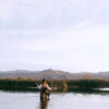 A female angler wearing waders stands knee-deep in a calm river, fly fishing Idaho. Tall grass and reeds line the bank, with mountains visible in the background under a mostly clear sky.