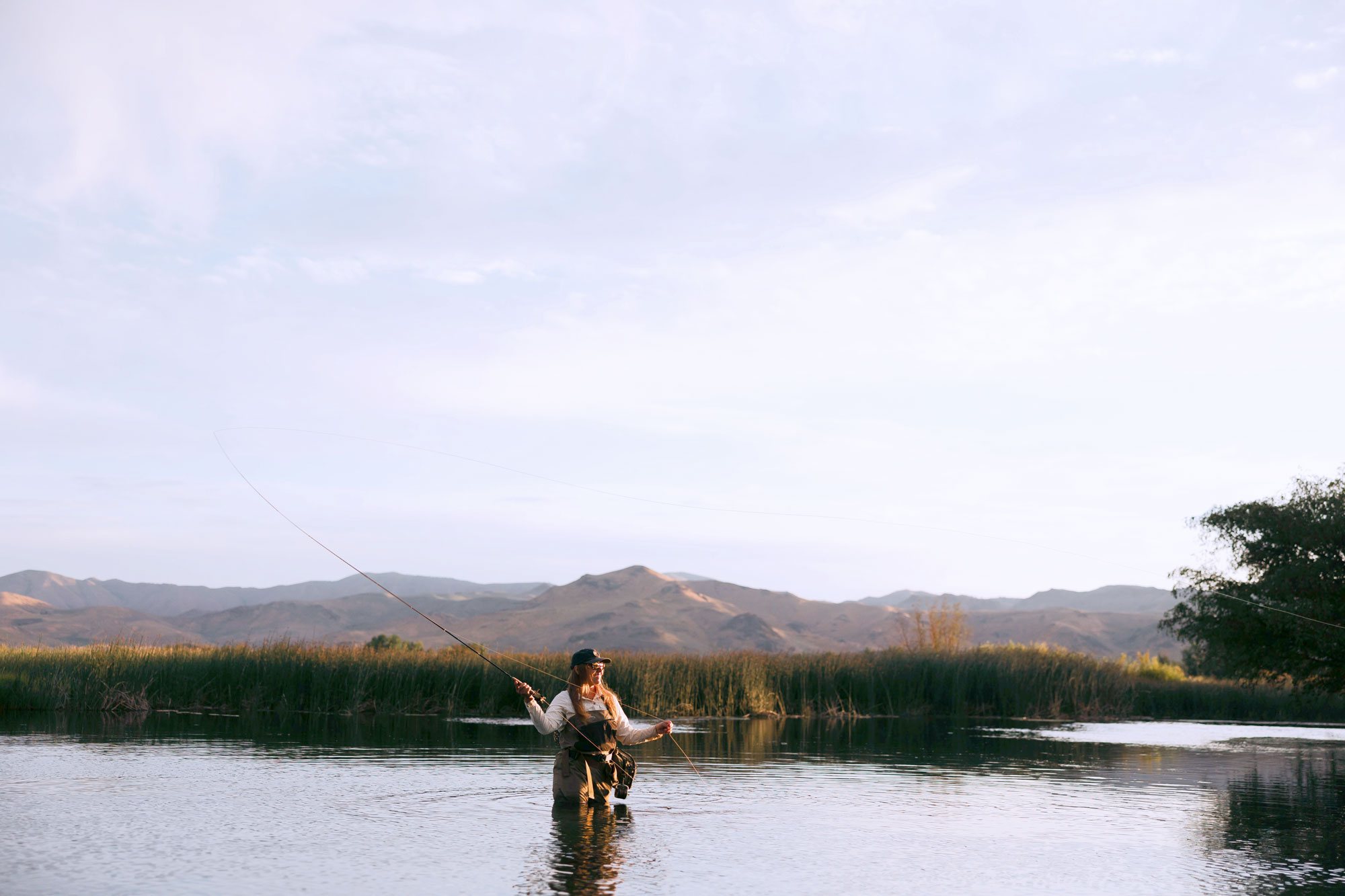 A female angler wearing waders stands knee-deep in a calm river, fly fishing Idaho. Tall grass and reeds line the bank, with mountains visible in the background under a mostly clear sky.