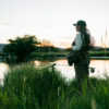 A person dressed in fishing gear stands in tall grass by a calm river at sunset, holding a fishing rod and net—capturing the peaceful spirit of Sun Valley Fly Fishing with trees and hills visible in the background.