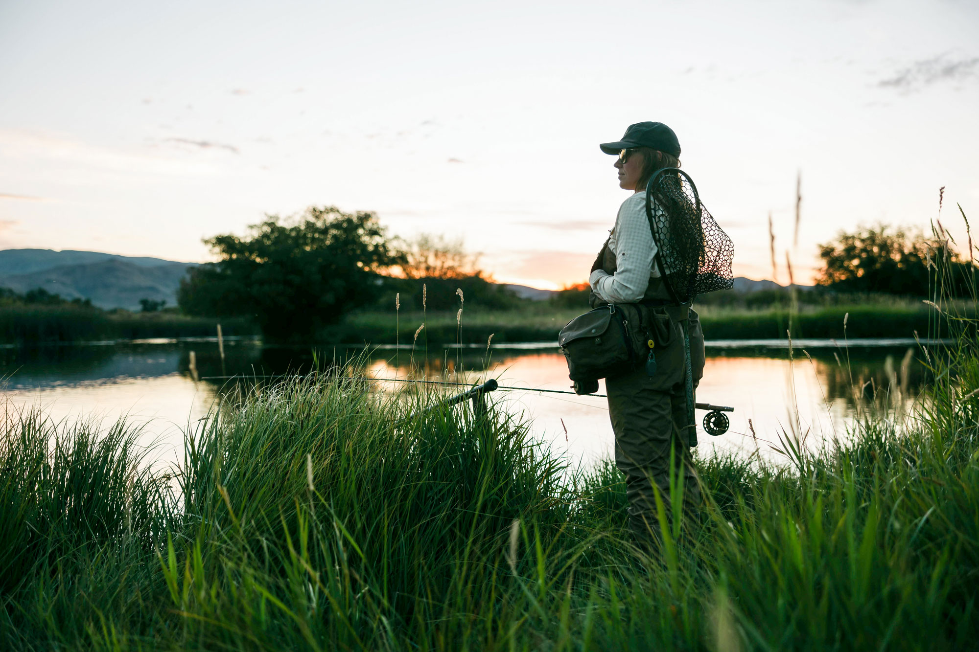 A person dressed in fishing gear stands in tall grass by a calm river at sunset, holding a fishing rod and net—capturing the peaceful spirit of Sun Valley Fly Fishing with trees and hills visible in the background.