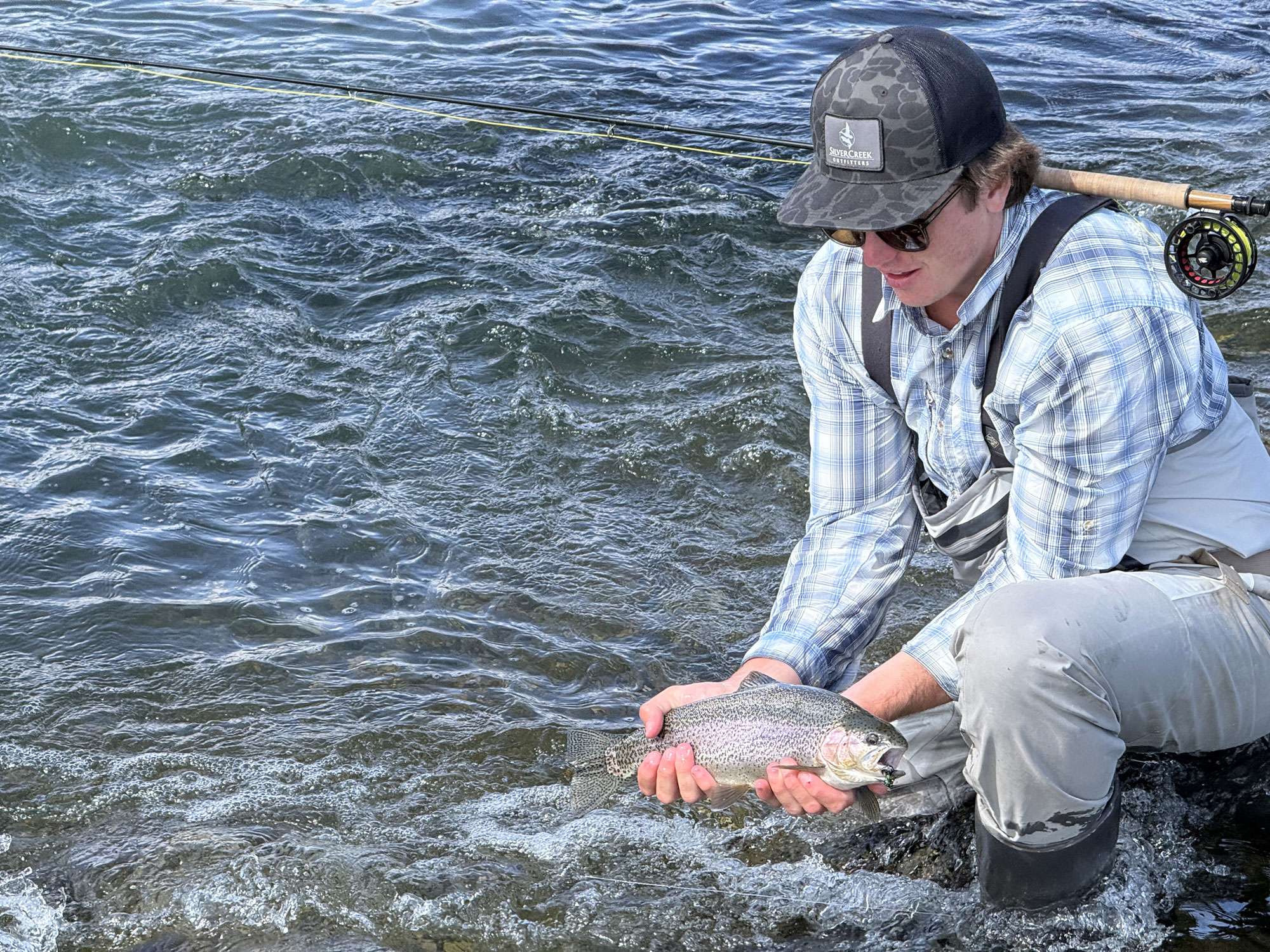 A person in fishing gear crouches by a river in Sun Valley, holding a large trout with both hands just above the water’s surface. A fishing rod rests on their shoulder.