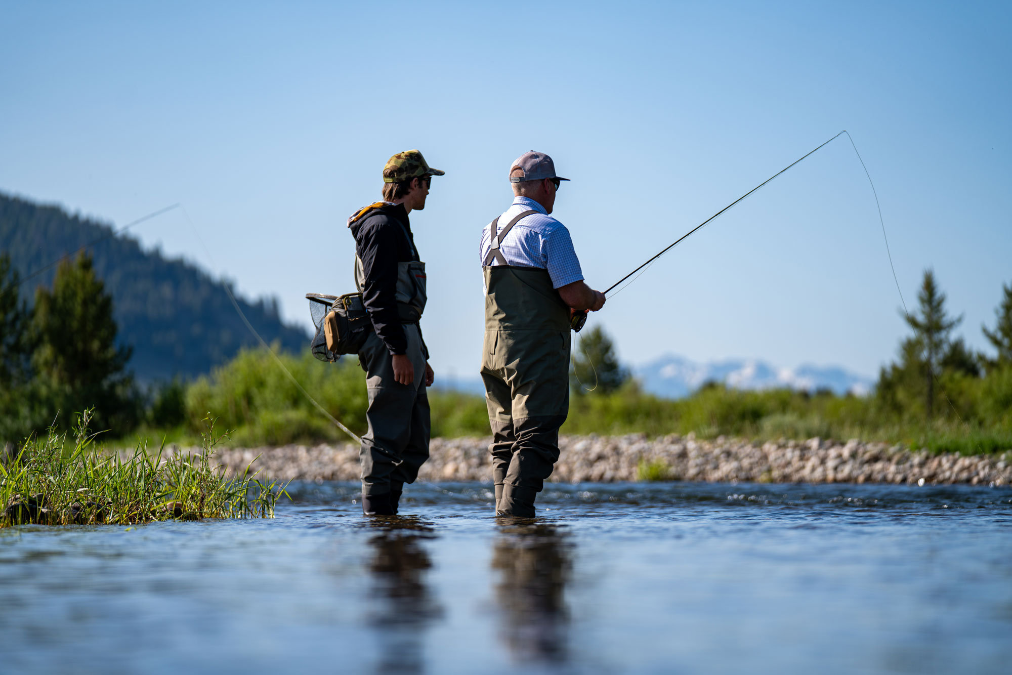 Two people in waders stand in a shallow river fly fishing, surrounded by greenery and distant mountains under a clear blue sky—an ideal scene for Sun Valley Fly Fishing enthusiasts seeking the perfect Fly Fishing Forecast.