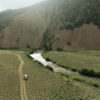 A white SUV drives on a dirt road through a grassy valley near Sun Valley Fly Fishing waters, beside a winding river, with forested hills and rocky slopes in the background under a cloudy sky.