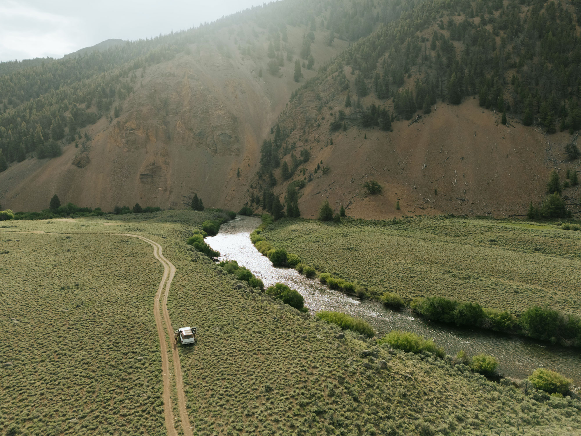 A white SUV drives on a dirt road through a grassy valley near Sun Valley Fly Fishing waters, beside a winding river, with forested hills and rocky slopes in the background under a cloudy sky.