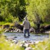 A person wearing a hat and outdoor clothing stands in a shallow, rocky stream while fly fishing, surrounded by lush green foliage—capturing the essence of Sun Valley Fly Fishing in Idaho.