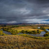 Photo: Arian Stevens. A winding river flows through a green, grassy plain under a dramatic, cloudy sky, perfect for Sun Valley Fly Fishing, with distant mountains on the horizon and patches of sunlight illuminating parts of the landscape.