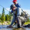 Two people enjoy fly fishing Idaho in a shallow river; one kneels to release a caught fish from a net, while the other stands behind, smiling. Trees and mountains rise in the background under a clear blue sky.