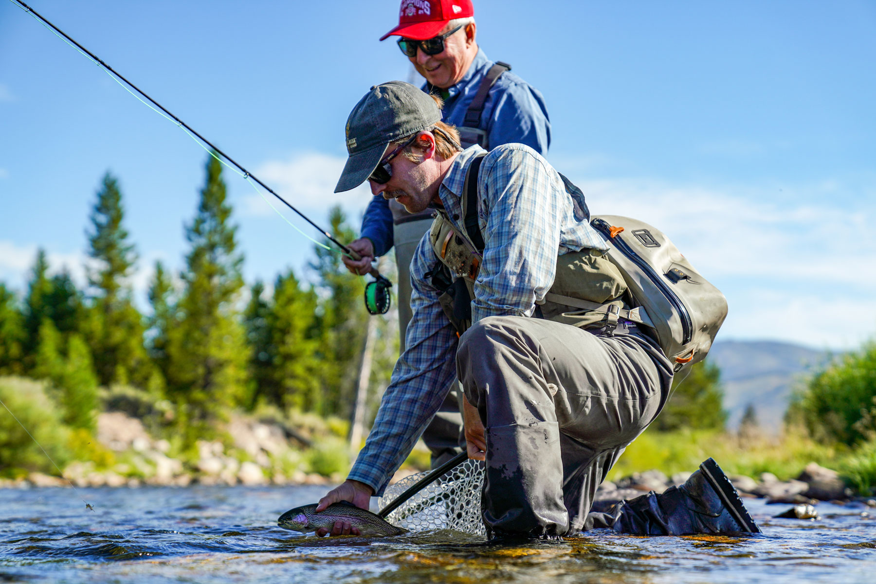 Two people enjoy fly fishing Idaho in a shallow river; one kneels to release a caught fish from a net, while the other stands behind, smiling. Trees and mountains rise in the background under a clear blue sky.