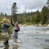 Two people stand in a shallow river, fly fishing Idaho style. One holds a fishing rod and net with a fish, smiling, while the other gives a high five. Trees and rocks line the riverbank under a cloudy sky.