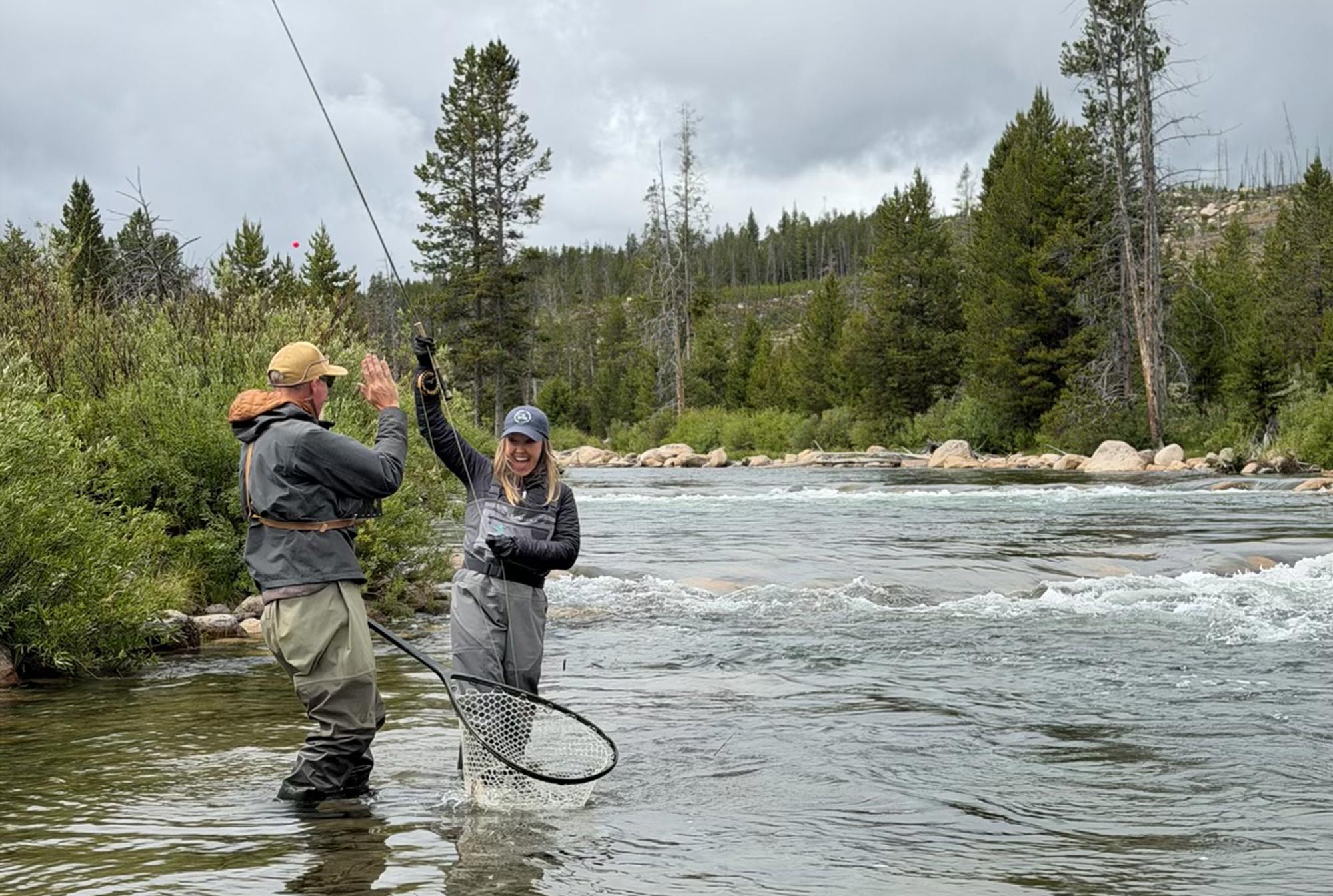 Two people stand in a shallow river, fly fishing Idaho style. One holds a fishing rod and net with a fish, smiling, while the other gives a high five. Trees and rocks line the riverbank under a cloudy sky.