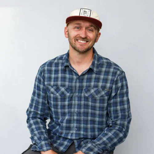 A man wearing a blue plaid shirt, gray pants, and a beige and maroon cap sits on a stool against a plain light background, smiling at the camera—ready for his next adventure as a Sun Valley Fly Fishing Guide.