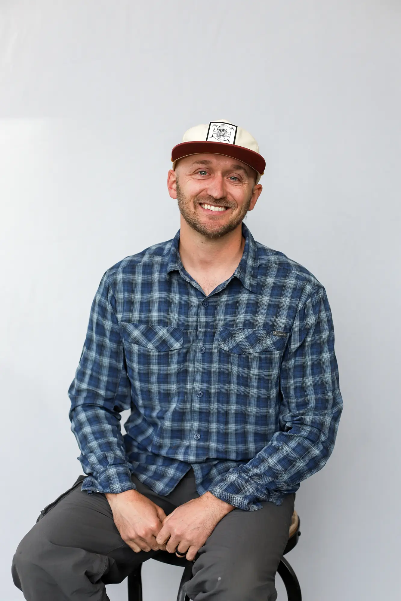 A man wearing a blue plaid shirt, gray pants, and a beige and maroon cap sits on a stool against a plain light background, smiling at the camera—ready for his next adventure as a Sun Valley Fly Fishing Guide.