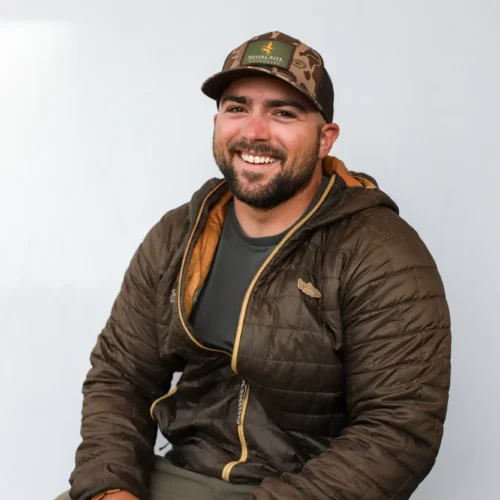 Lewis Hunt, a Certified Casting Instructor, smiles at the camera while sitting. He wears a brown puffer jacket, dark t-shirt, olive pants, and camo baseball cap, holding sunglasses in one hand against a plain light-colored background.