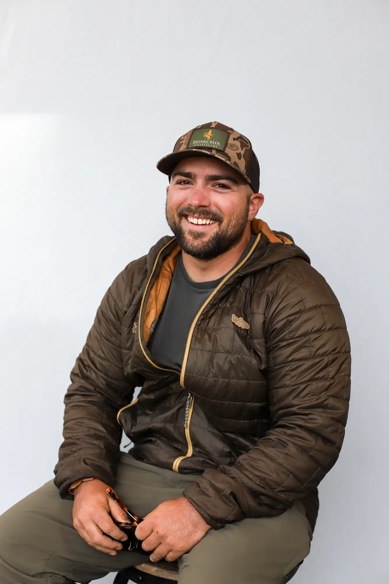 Lewis Hunt, a Certified Casting Instructor, smiles at the camera while sitting. He wears a brown puffer jacket, dark t-shirt, olive pants, and camo baseball cap, holding sunglasses in one hand against a plain light-colored background.
