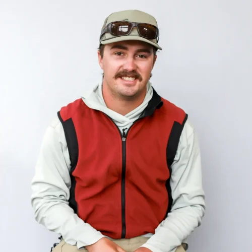 A man with a mustache wearing a light gray shirt, red and black vest, tan pants, a cap, and sunglasses sits on a stool against a plain light gray background.