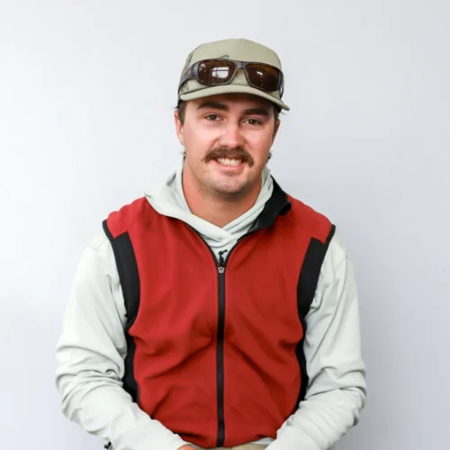 A man with a mustache wearing a light gray shirt, red and black vest, tan pants, a cap, and sunglasses sits on a stool against a plain light gray background.