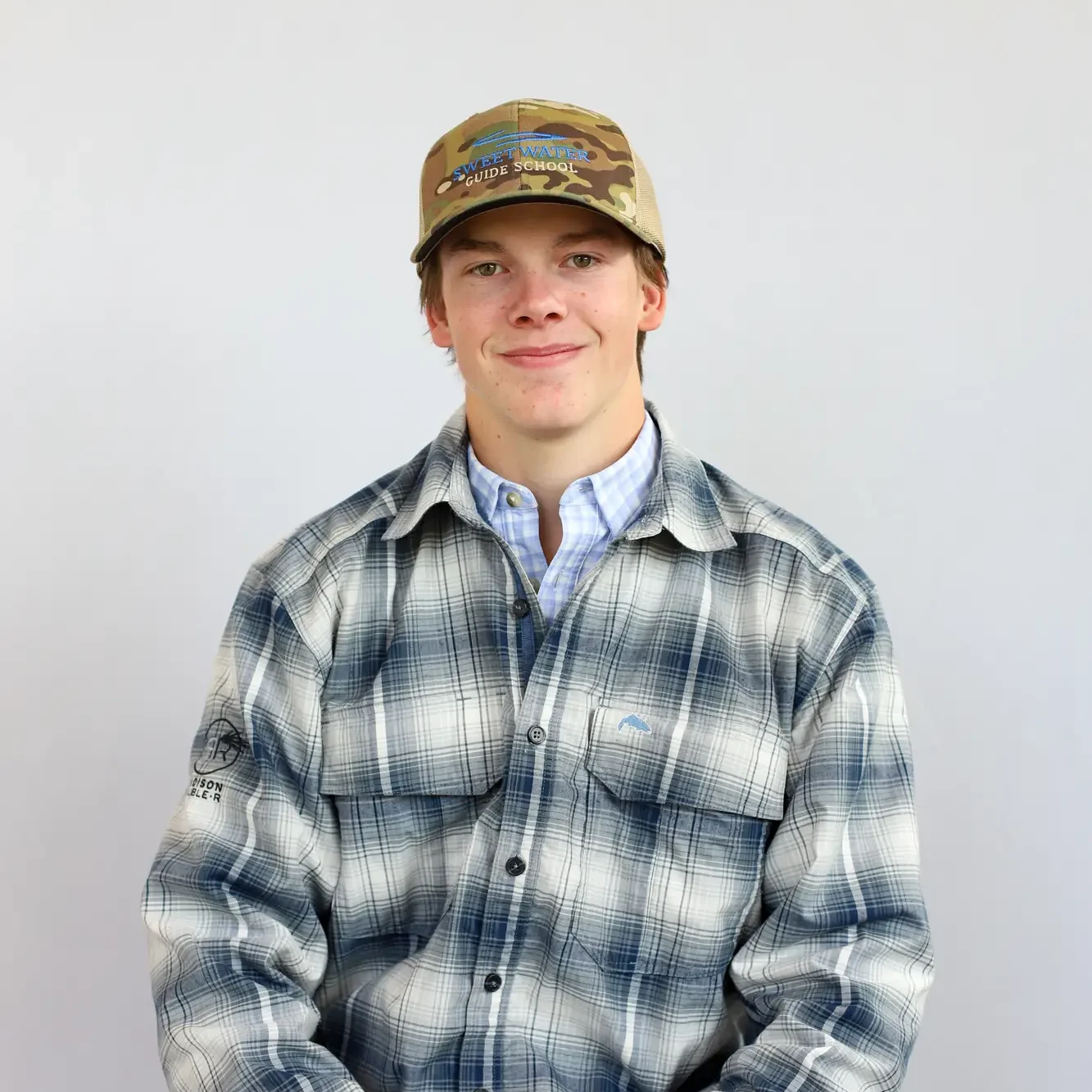 Jackson Gabbert, a Retail Associate, sits against a plain light background in a tan baseball cap, blue and white plaid button-up shirt, and gray pants, looking at the camera with a slight smile.