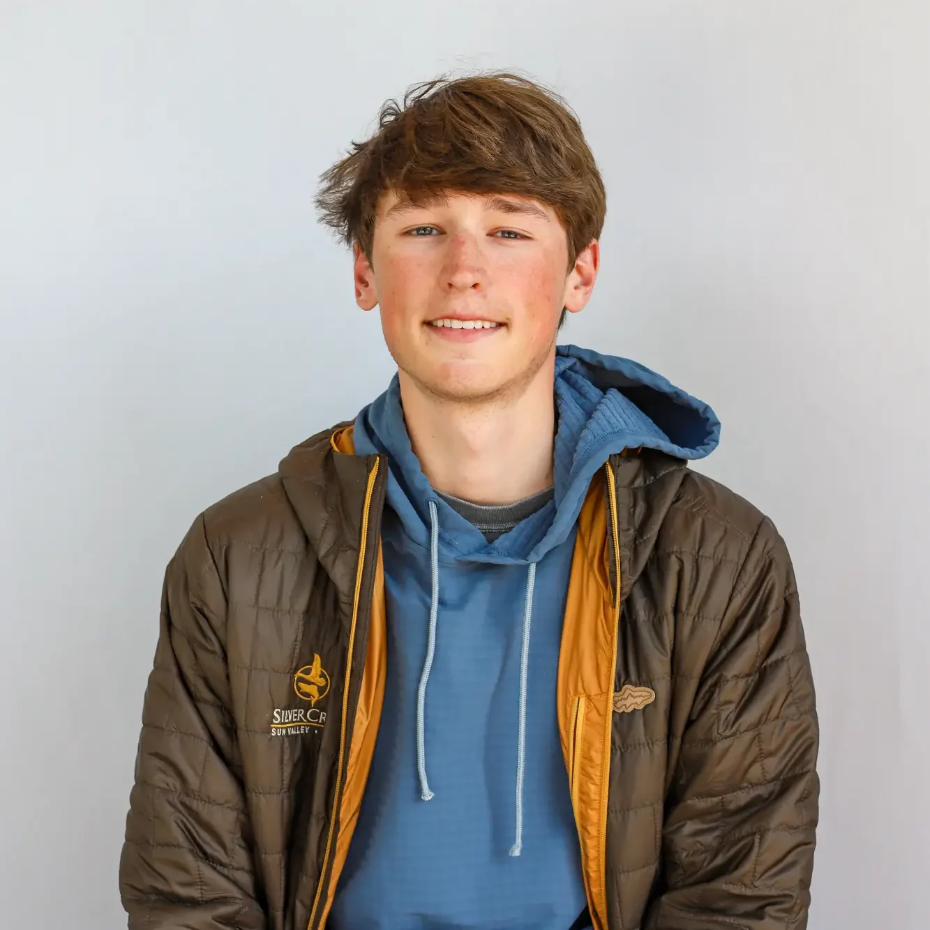 A young man with light brown hair sits facing the camera, wearing a brown puffer jacket over a blue hoodie. He is smiling softly against a plain light-colored background, embodying the relaxed confidence of a retail associate at Silver Creek Outfitters