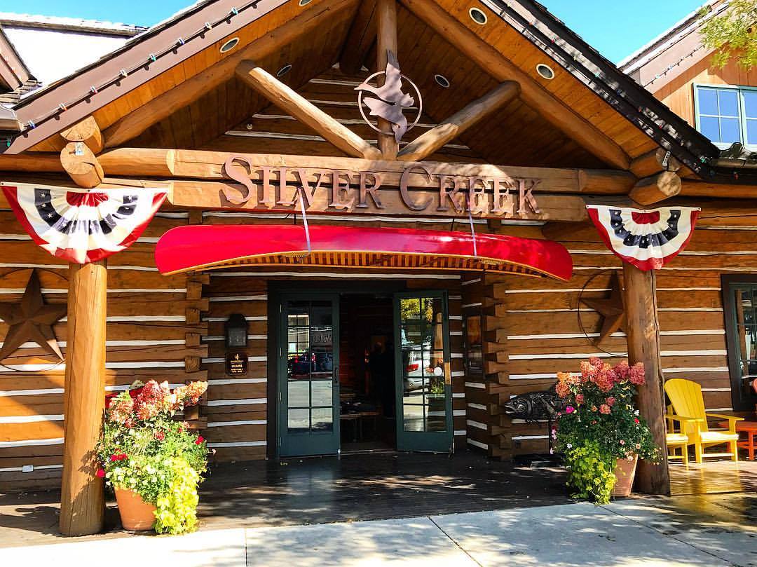 The entrance of Silver Creek, a log cabin-style building with a red canoe above the doors, patriotic bunting, wooden benches, potted plants, and rustic decor under a sunny blue sky.