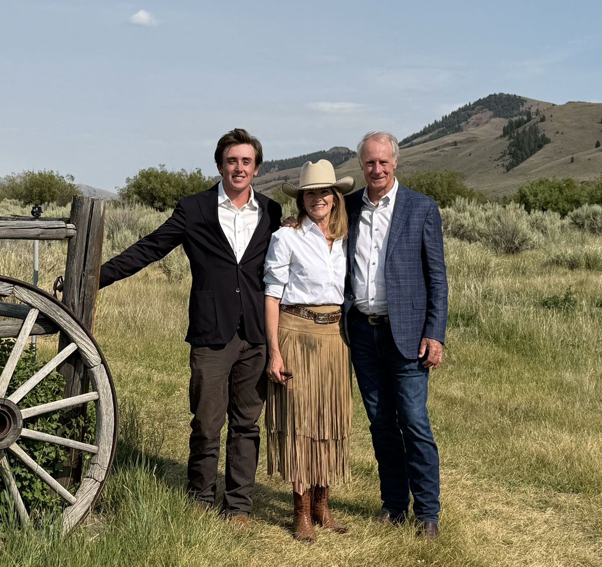 Three people stand together smiling in a grassy field with hills and shrubs in the background. One person wears a suit, another wears a cowboy hat, white shirt, and fringe skirt, and the third wears a blazer and jeans. A wagon wheel is on the left.